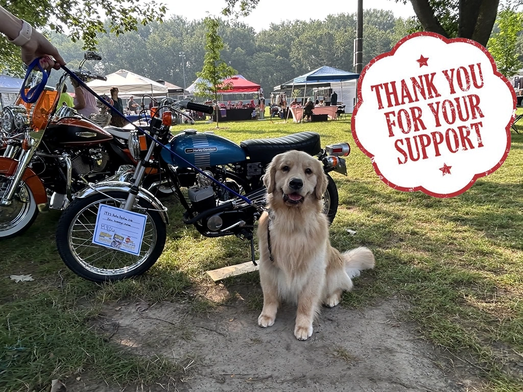 Golden retriever sits beside vintage motorcycles at the Flags & Honor of Minnesota Car Show with vendor tents in the background.
