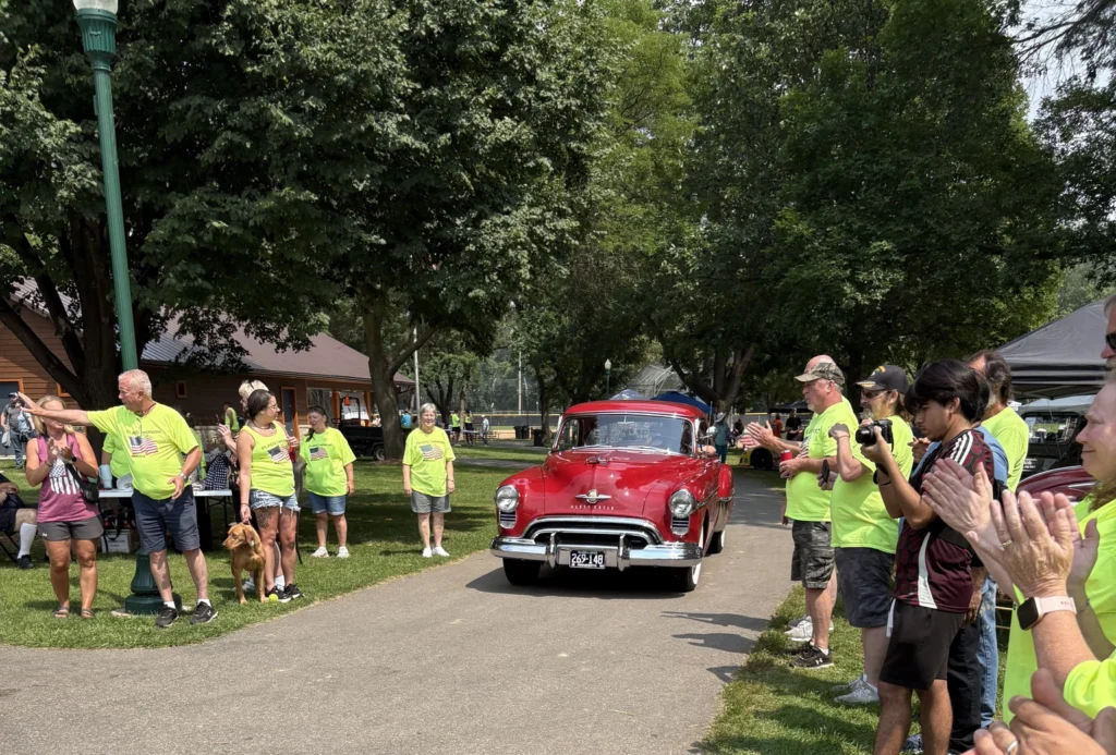 Spectators and Flags & Honor of Minnesota volunteers applaud as a classic red car drives through the annual Car Show.