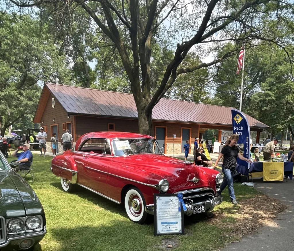 Classic red Oldsmobile on display at the Flags & Honor of Minnesota Car Show with the pavilion building in the background.